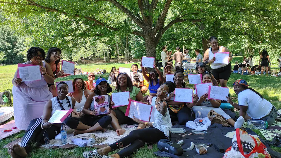 A big group of women sitting on blankets in a park holding pink papers